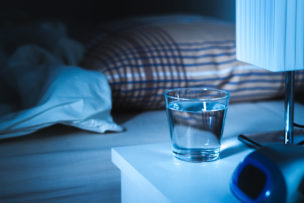 Sleeping water on nightstand next to a glass of water. White tablet on table and bed in bedroom.
