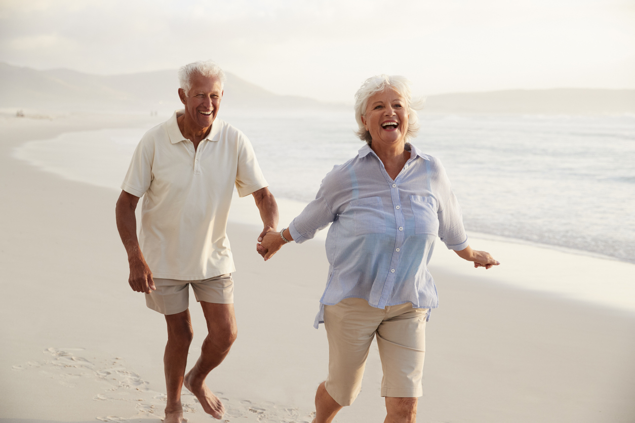 Senior Retired Couple Running Along Beach Hand In Hand Together