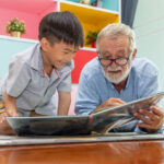 Happy boy grandson reading book with old senior man grandfather at home
